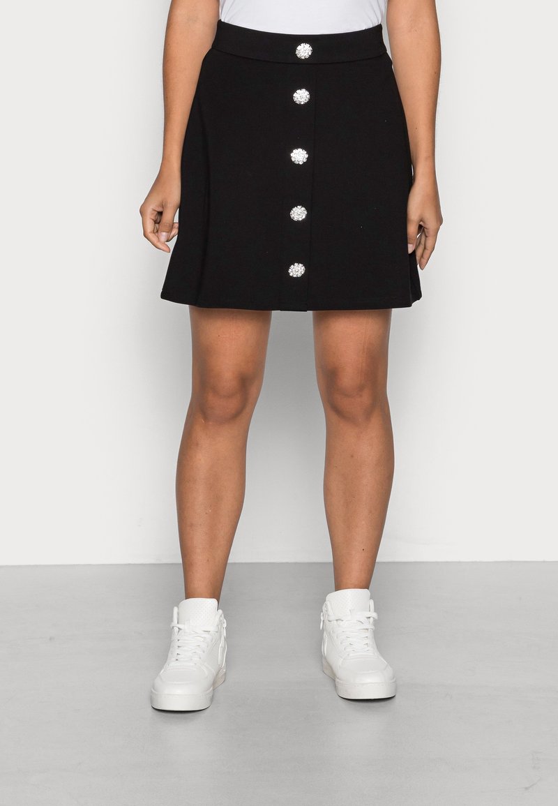Femme portant une jupe noire avec cinq grands boutons décoratifs à l’avant et des baskets blanches montantes, debout sur un sol gris.