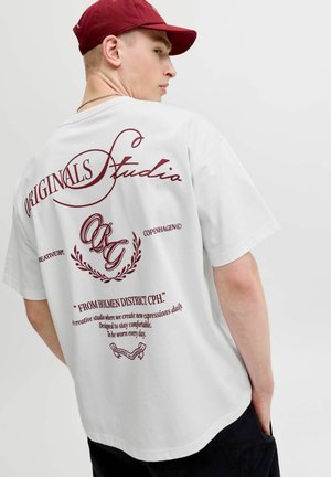 Jeune homme portant un t-shirt blanc avec un texte et des graphismes rouges dans le dos et une casquette rouge, regardant par-dessus son épaule sur un fond uni.