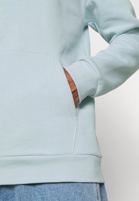 Sudadera azul claro con textura suave y bolsillo tipo canguro. Cuenta con dobladillo y puños acanalados; diseño casual con costuras mínimas.