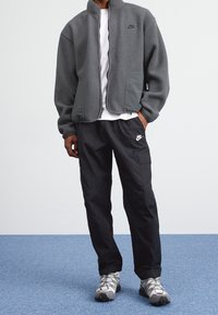 Giacca in pile grigia con zip e colletto alto, abbinata a pantaloni cargo neri con tasca laterale e logo bianco.