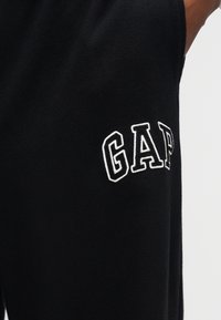 Μαύρο ύφασμα με λευκά γράμματα "GAP" με περίγραμμα, κεντημένα στην πλευρά ενός μανικιού ή ποδιού παντελονιού, που φοριέται από ένα άτομο.