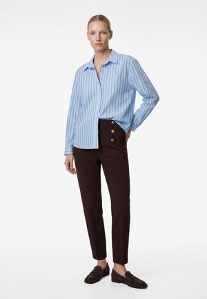 Femme portant une chemise rayée bleu clair à boutons, un pantalon marron foncé avec des boutons dorés, et des mocassins marron foncé, debout avec la main sur la hanche.