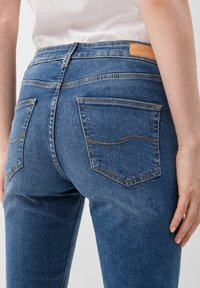 Modre denim hlače z srednjim pasom, dvema zadnjima žepoma z rjavim šivom in rjavim usnjenim označevalcem na zadnjem pasu.
