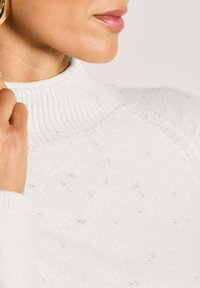 Frau, die einen weiß melierten Strickpullover mit hohem Rippkragen und goldenen Ohrhängern trägt, während sie mit der Hand den Kragen nahe des Halses berührt.