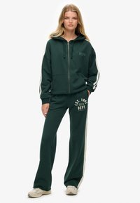 Groene rits-hoodie met een trekkoord, gecombineerd met bijpassende joggingbroek met een logo en contrasterende zijstrepen. Materiaal lijkt zacht en comfortabel.