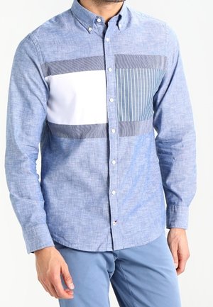 Uomo che indossa una camicia azzurro chiaro con pannelli orizzontali bianchi e blu scuro e righe verticali sulla parte superiore del petto, abbinata a pantaloni azzurri.