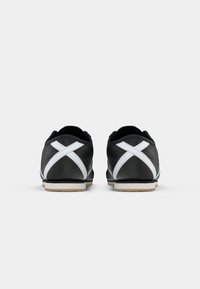 Pár černých sneakers s bílým křížovým pruhem na zádech a béžovými podrážkami, z pohledu zezadu na jednobarevném pozadí.