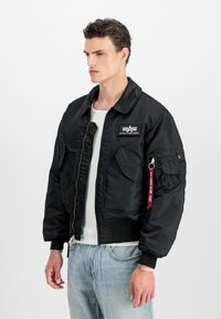 Mand i sort Alpha Industries bomberjakke med forlommer, "Remove Before Flight" mærke på ærmet, hvid skjorte og lyseblå jeans.