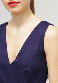 Žena v bezramínkových šatech tmavě modré barvy s hlubokým V výstřihem a jasně červenými rtěnkami, zobrazená od ramen po střed hrudníku na jednobarevném pozadí.