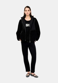 Giacca nera con zip in una texture morbida, indossata sopra una maglietta nera con scritte bianche, abbinata a pantaloni neri e ballerine con stampa.