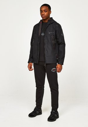 Uomo che indossa una giacca nera con cappuccio, una T-shirt nera con grafica, pantaloni della tuta neri con toppa e sneaker nere, in piedi contro uno sfondo chiaro e uniforme.