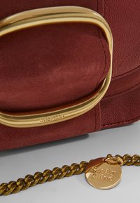 Röd läderväska med en guldfärgad rektangulär hårdvaruaccents och en kedjestropp med en rund tagg, präglad med varumärkesnamnet.
