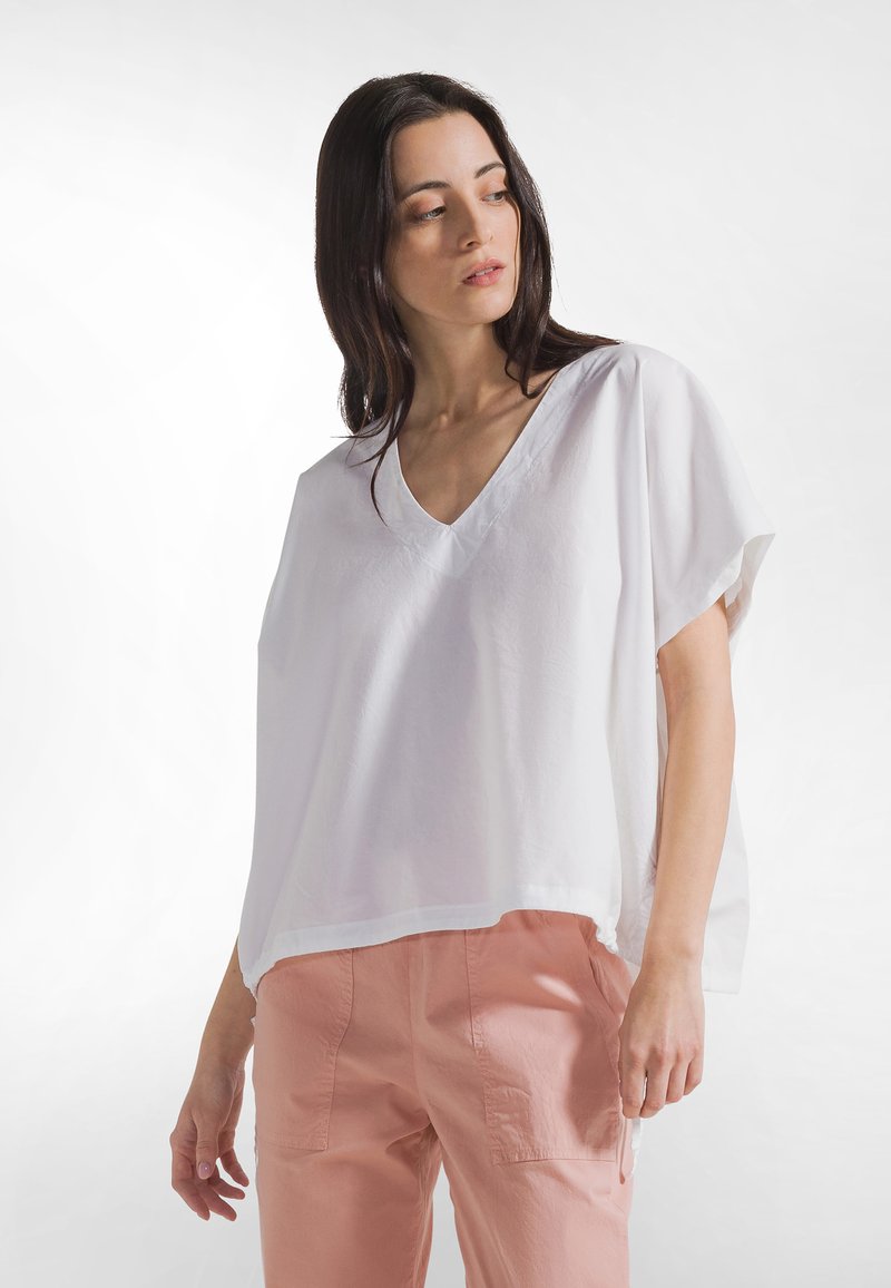 Blusa blanca de escote en V, de corte holgado y confeccionada en tela ligera. Cuenta con mangas cortas y dobladillo curvado, combinada con pantalones rosa claro.