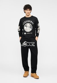 Μαύρο πουκάμισο με μακριά μανίκια και γραφικά, και μαύρο φόρμα παντελόνι με λευκό σχέδιο καρτούν σκύλου, συνδυασμένα με μπεζ μπότες.