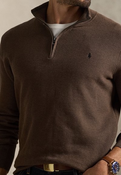 Rudas megztinis su apykakle, užsegama užtrauktuku. Turi struktūruotą raštą ir mažą logotipą krūtinės srityje. Dėvimas ant balto marškinio.