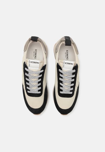 Zapatillas deportivas beige y negras con una parte superior de ante y cuero, cordones blancos con un diseño estampado y una suela de goma color beige.