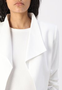 Bílý blejzr s velkým límcem, hladkou texturou a nařasenými rukávy; vrstvený přes bílý přiléhavý top. Čistý, minimalistický design.