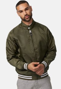Giacca bomber verde oliva con colletto alzato, chiusura con zip e polsini e orlo a costine bianchi. Tessuto liscio e lucido con tasche laterali.