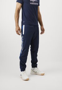 Calças de treino navy com detalhes brancos e azuis, apresentando um grande logotipo "LACOSTE" na lateral e punhos elásticos nos tornozelos.