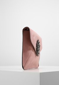 Bolso de mano de ante rosa con cierre de solapa, que presenta un adorno decorativo de tono plateado en la parte delantera. Textura suave y forma estructurada.