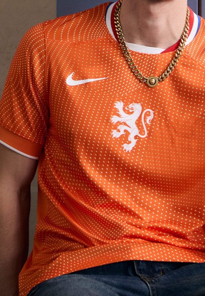 Maillot Nike orange vif avec motif à points blancs, arborant un logo de lion blanc sur la poitrine et un col rond côtelé. Chaîne en or visible.