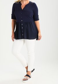 Haut bleu marine à boutons avec manches trois-quarts et taille froncée, associé à un pantalon blanc et des sandales noires avec des accents argentés.