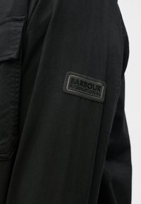 Fekete dzseki sima textúrával, a bal ujján dombornyomott "BARBOUR INTERNATIONAL" felirattal ellátott folttal.