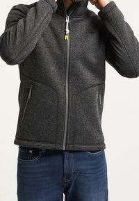 Šedá bunda na zip z texturované látky se dvěma bočními kapsami, přiléhavým střihem a stojáčkovým límcem, s kontrastním přívěskem na zipu.