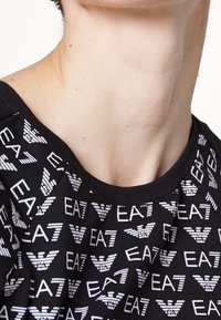 Svart bomulls-T-shirt med vid halsringning, prydd med ett upprepat vitt logomönster. Mjuk textur med korta ärmar och en avslappnad passform.