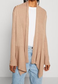 Kremový žebrovaný cardigan s otevřeným předním dílem a dlouhými rukávy, který má strukturovaný design, měkký pletený materiál a splývavý asymetrický lem.