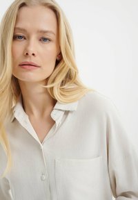 Camisa de lino beige claro con cuello abotonado, bolsillo en el pecho y corte relajado. Textura sutil visible en la tela.