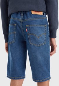 Blue denim shorts with a relaxed fit, featuring two back pockets, yellow stitching, and a leather patch on the waistband.