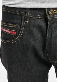 Mustad denim püksid kontrastsete kollaste õmblustega, punase sildiga tagaosal, ning metallist nööpkinnitusega vöökohal.