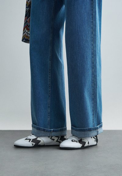 Άσπρα και μαύρα παπούτσια με γεωμετρικό σχέδιο και σπορ σχεδίαση, φορεμένα με μπλε, φαρδιά τζιν παντελόνια που διαθέτουν διπλωμένο στρίφωμα.