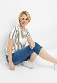 Camicia a righe con maniche corte in crema e nero, abbinata a jeans blu taglio cropped e sneaker bianche. Posizione seduta con uno sfondo neutro.