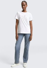 Weißes Baumwoll-T-Shirt mit lockerem Schnitt, versehen mit "5th Ave Club" in grauer, geschwungener Schrift. Kombiniert mit blauen Jeans im geraden Schnitt und weißen Sneakers.