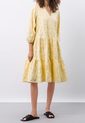 Frau trägt ein gelbes Midikleid mit Blumenstickerei, dreiviertel Puffärmeln und schwarzen flachen Sandalen, steht auf grauem Boden.