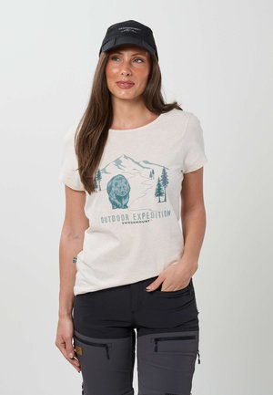 Frau trägt eine schwarze Kappe und ein beiges T-Shirt mit Berg-, Bären- und Baum-Grafik mit der Aufschrift "Outdoor Expedition Swedemount" sowie schwarze Cargohosen.