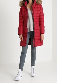 Röd puffjacka med en pälsfodrad huva, quiltad textur, lång längd, tillsammans med en grå topp och grå jeans, avslutad med vita sneakers.