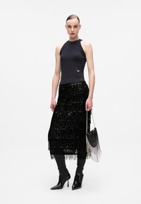 Top noir sans manches avec des accents scintillants, associé à une longue jupe à franges en sequins noirs. Le mannequin tient un petit sac noir avec un détail en chaîne.