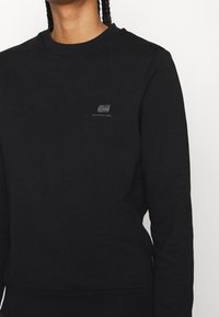 Sweat-shirt en coton noir avec un col rond, arborant un petit logo sur la poitrine. Texture lisse et coupe ajustée avec des poignets côtelés.