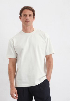 Maglietta bianca in cotone con scollo rotondo, maniche corte e vestibilità rilassata, indossata con pantaloni scuri su uno sfondo neutro.