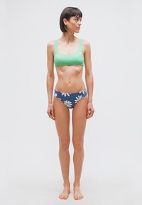 Femme debout pieds nus portant un crop top vert et un bas de bikini bleu avec de grandes impressions de marguerites blanches, regardant vers sa gauche sur un fond uni.