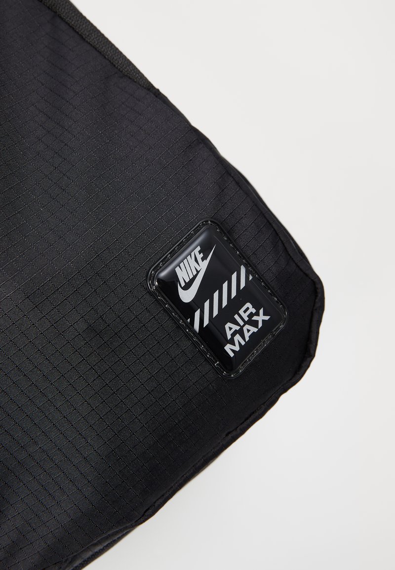 Schwarze, strukturierte Stofftasche mit einem rechteckigen Nike Air Max Logo-Patch, das weiße Schriftzüge und ein gestreiftes Designelement aufweist.