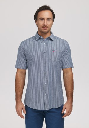 Mężczyzna ubrany w niebiesko-białą koszulę na krótki rękaw z pionowymi paskami i niebieskie spodnie, stojący na tle jednolitego tła.