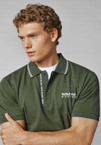 Olivgrön polo med vita detaljer och branding. Har en krage, tre knappar och korta ärmar. Tyget verkar slätt och strukturerat.