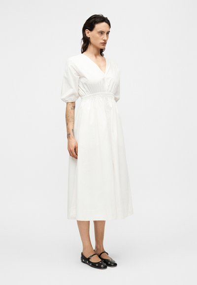 Λευκό φόρεμα μέσου μήκους με V-λαιμόκοψη και ελαστική μέση που μαζεύει. Μανίκια καμπάνα και λεία υφή. Μαύρα επίπεδα παπούτσια με διακοσμητικές λεπτομέρειες.