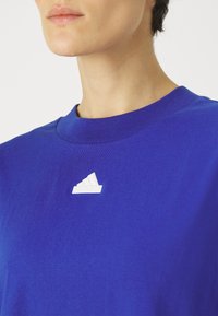 Modré bavlněné tričko s kulatým výstřihem a krátkými rukávy. Na hrudi se nachází bílé logo Adidas. Hladká textura a ležérní design.