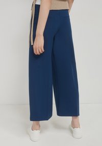 Persona che indossa pantaloni larghi blu, una maglietta beige e sneakers bianche, in piedi davanti a uno sfondo bianco uniforme.