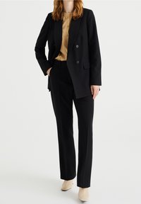 Blazer negro de doble botonadura con solapa muesca, llevado sobre una camisa dorada de botones. Combinado con pantalones negros a medida y botas de tobillo beige.